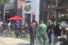 People line up outside a storefront with a Teenage Mutant Ninja Turtles Pizzeria sign and a red umbrella on a city street.