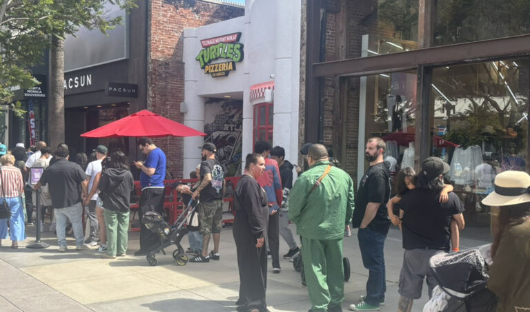 People line up outside a storefront with a Teenage Mutant Ninja Turtles Pizzeria sign and a red umbrella on a city street.