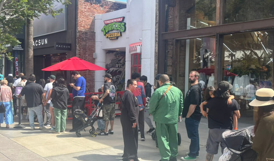 People line up outside a storefront with a Teenage Mutant Ninja Turtles Pizzeria sign and a red umbrella on a city street.