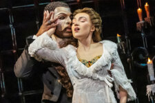 Masked man (Phantom) in a half-face mask holds Christine as she leans back on a candlelit stage.