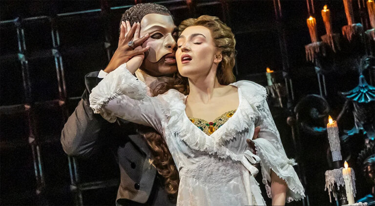 Masked man (Phantom) in a half-face mask holds Christine as she leans back on a candlelit stage.