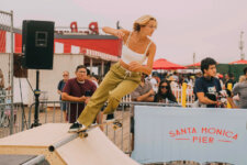 Skateboarder in a white tank top and khaki pants grinds a metal pipe on a wooden ramp at Santa Monica Pier, with spectators in the background.