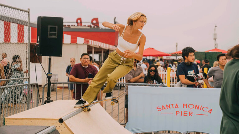 Skateboarder in a white tank top and khaki pants grinds a metal pipe on a wooden ramp at Santa Monica Pier, with spectators in the background.