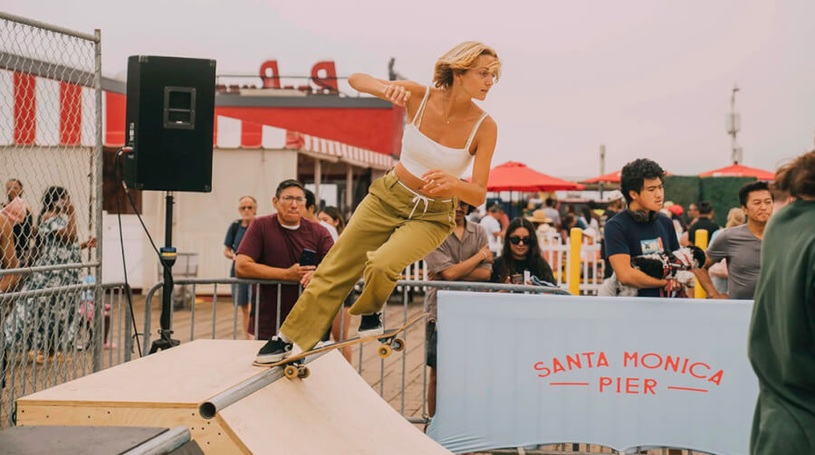 Skateboarder in a white tank top and khaki pants grinds a metal pipe on a wooden ramp at Santa Monica Pier, with spectators in the background.