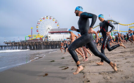 Group of swimmers in wetsuits and blue swim caps sprinting into the ocean from a sandy beach beside a boardwalk with a large Ferris wheel in the background.