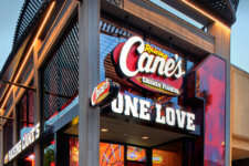 Exterior of a Raising Cane's restaurant with large illuminated 'Cane's' logo and 'ONE LOVE' sign on the storefront.