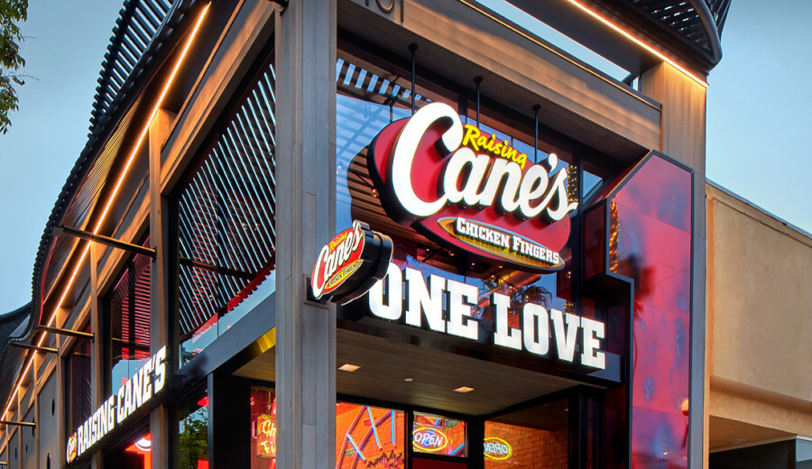 Exterior of a Raising Cane's restaurant with large illuminated 'Cane's' logo and 'ONE LOVE' sign on the storefront.