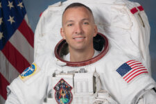 Astronaut in a white spacesuit, smiling, with an American flag patch on the sleeve and the US flag in the background.