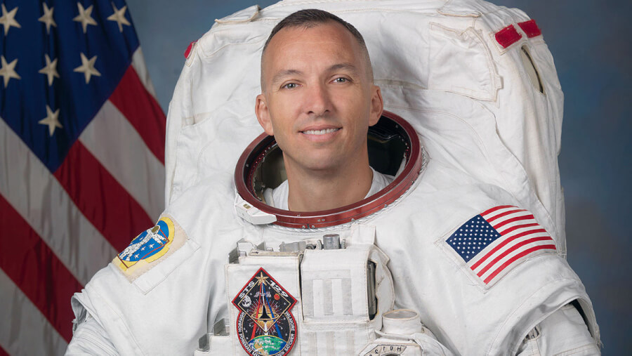Astronaut in a white spacesuit, smiling, with an American flag patch on the sleeve and the US flag in the background.