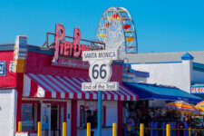 Route 66 End of the Trail sign in front of a colorful boardwalk shop with a ferris wheel in the background on Santa Monica Pier, blue sky above.