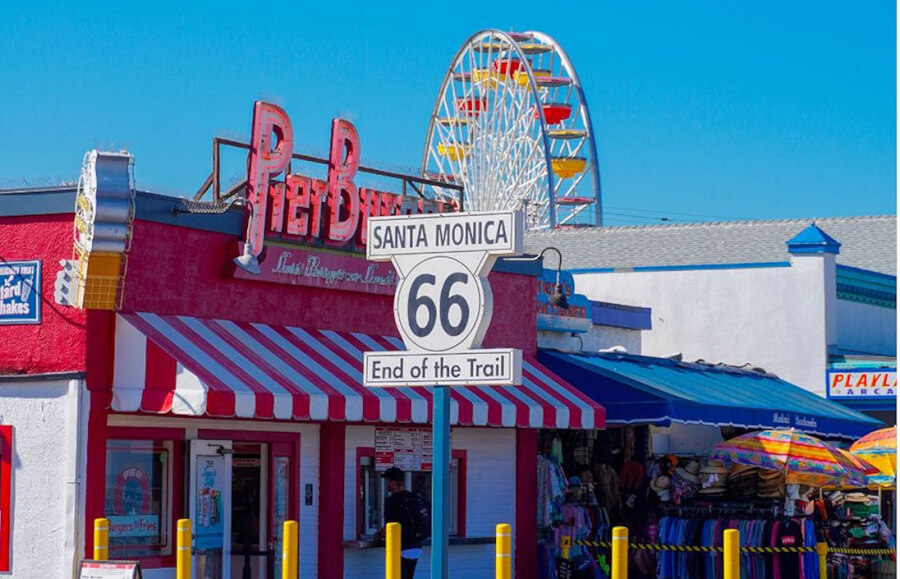 Route 66 End of the Trail sign in front of a colorful boardwalk shop with a ferris wheel in the background on Santa Monica Pier, blue sky above.