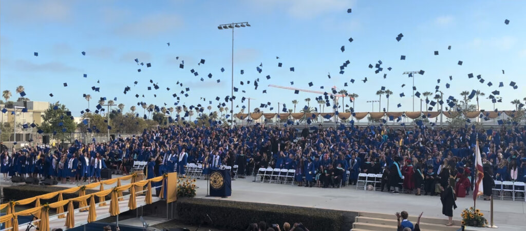 Crowd of graduates in blue gowns toss their caps in the air at an outdoor commencement, with a decorated stage and palm trees in the background.