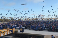 Crowd of graduates in blue gowns toss their caps in the air at an outdoor commencement, with a decorated stage and palm trees in the background.