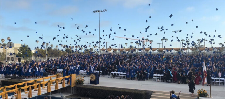 Crowd of graduates in blue gowns toss their caps in the air at an outdoor commencement, with a decorated stage and palm trees in the background.