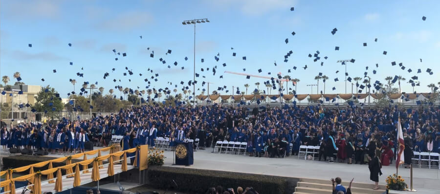 Crowd of graduates in blue gowns toss their caps in the air at an outdoor commencement, with a decorated stage and palm trees in the background.