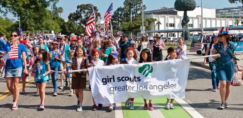 Group of Girl Scouts marching in a parade, holding a banner that reads 'girl scouts greater los angeles' with American flags in the crowd behind them.