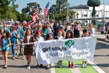 Group of Girl Scouts marching in a parade, holding a banner that reads 'girl scouts greater los angeles' with American flags in the crowd behind them.