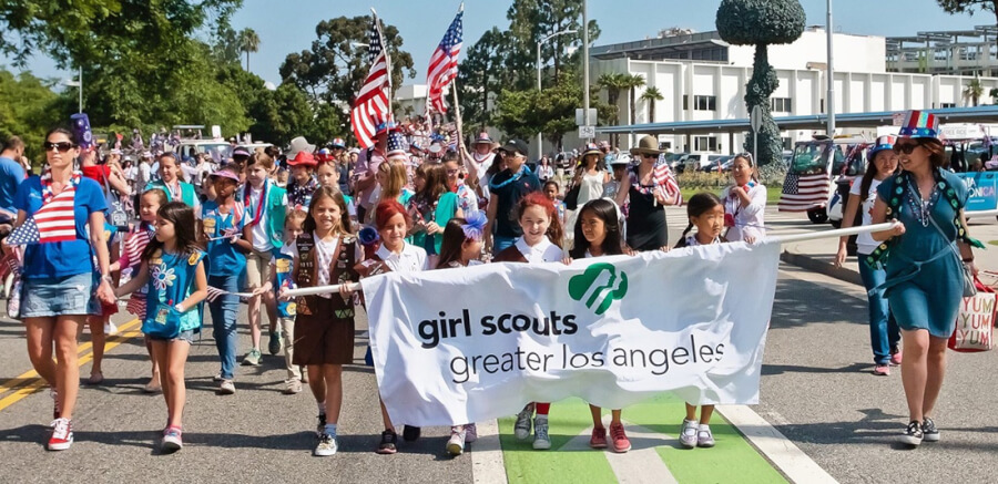 Group of Girl Scouts marching in a parade, holding a banner that reads 'girl scouts greater los angeles' with American flags in the crowd behind them.
