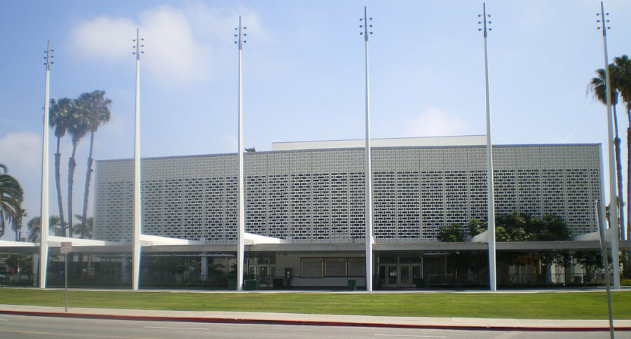 Public building with a perforated brick facade, seven flagpoles in front, and palm trees on a manicured lawn.