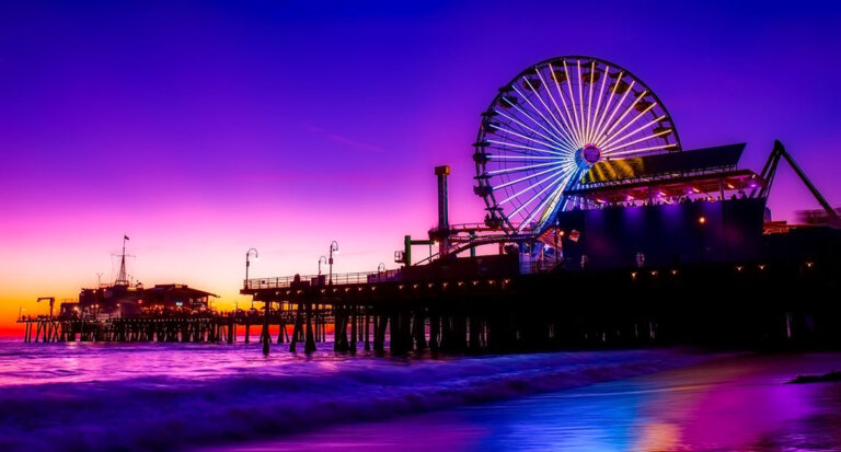 Colorful sunset over Santa Monica pier with a large illuminated Ferris wheel and calm waves reflecting the lights.