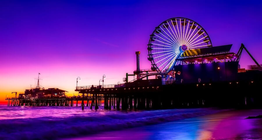 Colorful sunset over Santa Monica pier with a large illuminated Ferris wheel and calm waves reflecting the lights.
