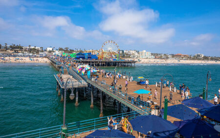 Pier extends into blue ocean with a ferris wheel and boardwalk crowded with people at a sunny beach town.