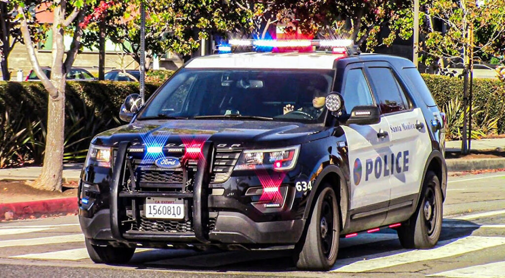 Police SUV with flashing red and blue lights driving on a street; Santa Monica Police markings on the side (POLICE).