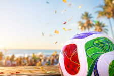 Close-up of a colorful FIFA World Cup soccer ball on a wooden table with a beach party, confetti in the air and palm trees in the background at sunset
