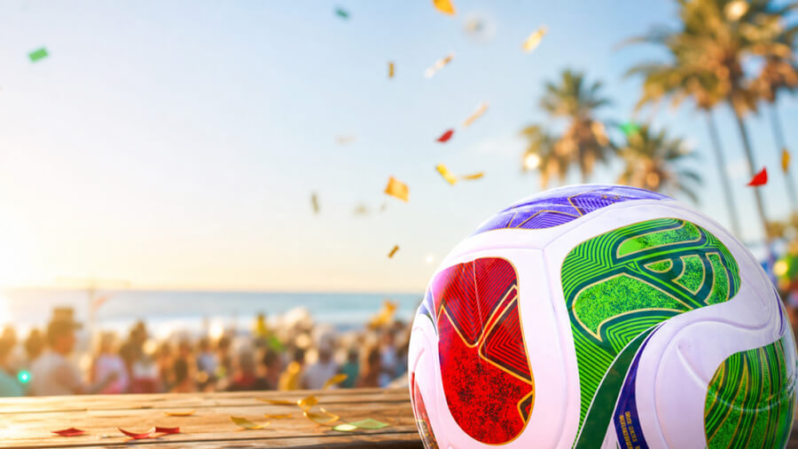 Close-up of a colorful FIFA World Cup soccer ball on a wooden table with a beach party, confetti in the air and palm trees in the background at sunset
