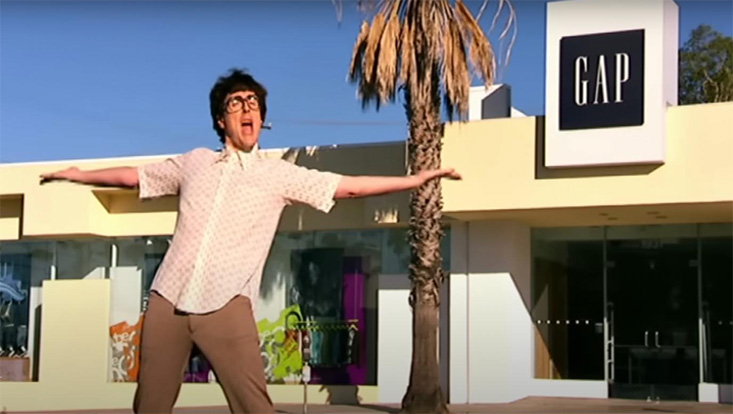 Young man with glasses dancing with arms outstretched in front of a GAP storefront on a sunny day.