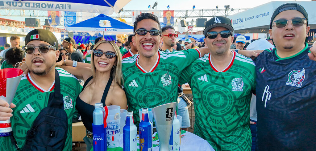 Group of smiling fans wearing green Mexico soccer jerseys at a crowded outdoor party, with drinks on the table and camaraderie visible.