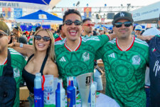 Group of smiling fans wearing green Mexico soccer jerseys at a crowded outdoor party, with drinks on the table and camaraderie visible.