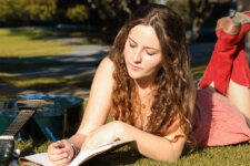 Young woman lying on grass in a park, writing in a notebook with a guitar beside her.