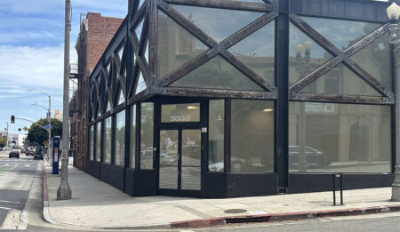 Corner storefront with large glass panels and black framing on a sunny urban street; door reads 3002 on the building corner.