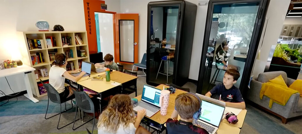 Students sit around tables with laptops in a bright, modern study space; some wear headphones and work intently on projects.