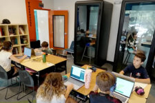 Students sit around tables with laptops in a bright, modern study space; some wear headphones and work intently on projects.