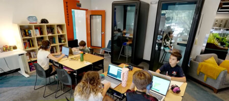 Students sit around tables with laptops in a bright, modern study space; some wear headphones and work intently on projects.