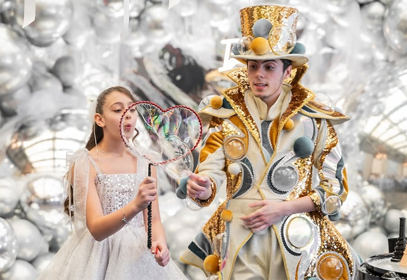 Girl in a sparkling silver dress holds a heart-shaped bubble wand next to a performer in a gold sequined costume at a festive event.