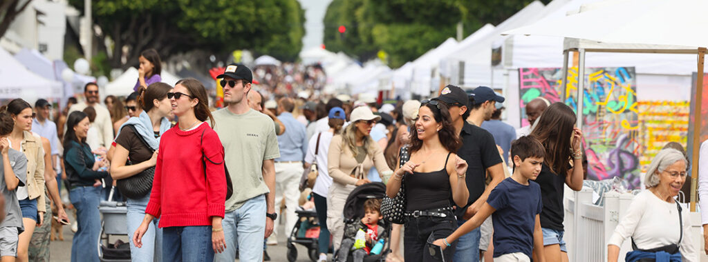 Crowded outdoor street market with white tents and people browsing stalls on a sunny day.