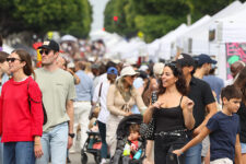 Crowded outdoor street market with white tents and people browsing stalls on a sunny day.