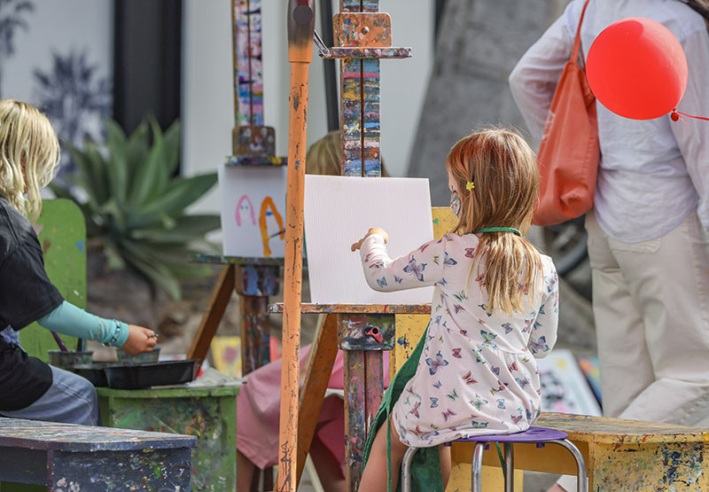 Young girl painting on a white canvas at a colorful outdoor art station, with a red balloon nearby.