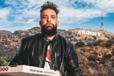 Bearded man in a black leather jacket holds a pizza box outdoors with the Hollywood sign in the hills behind him.