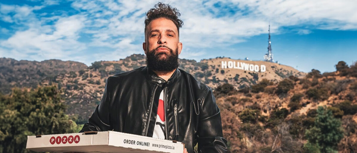 Bearded man in a black leather jacket holds a pizza box outdoors with the Hollywood sign in the hills behind him.