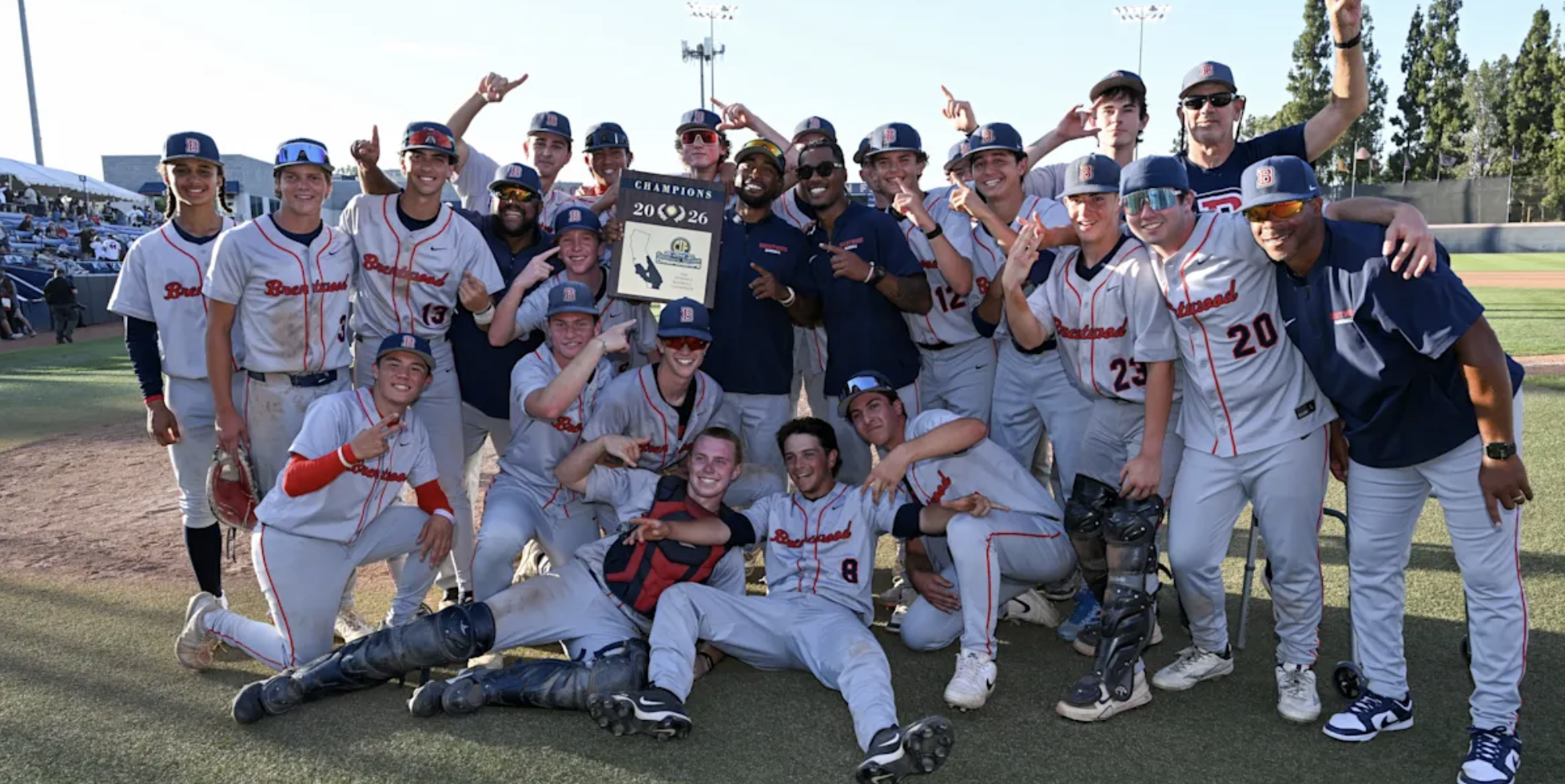 Baseball teammates celebrate winning the 2026 championship, posing with a plaque on the field.
