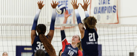 Indoor girls' volleyball game: a player in a red-blue jersey (#18) spikes the ball over the net while two defenders in dark jerseys (#34 and #21) jump to block, gym banners in the background.