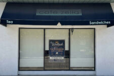 Front of a small café with a dark blue awning that says 'cafe zella', and signs 'Tea' and 'Sandwiches' on either side over a large glass window.