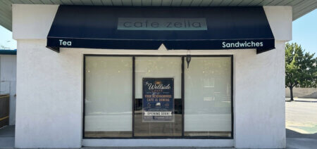 Front of a small café with a dark blue awning that says 'cafe zella', and signs 'Tea' and 'Sandwiches' on either side over a large glass window.