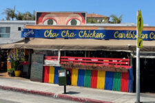 Colorful Caribbean restaurant with rainbow fencing and outdoor seating under a beige canopy; sign reads Cha Cha Chicken Caribbean Cuisine on a sunny street.