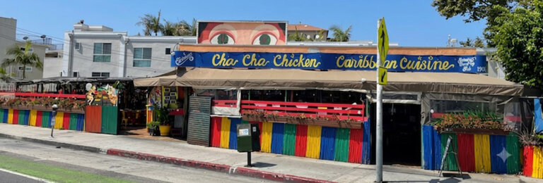 Colorful Caribbean restaurant with rainbow fencing and outdoor seating under a beige canopy; sign reads Cha Cha Chicken Caribbean Cuisine on a sunny street.