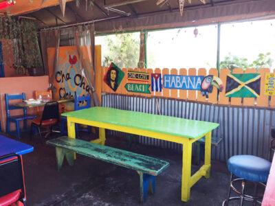 Colorful outdoor cafe with a lime-green table, yellow legs, and a weathered green bench along a fence with painted signs (Habana, Jamaica).